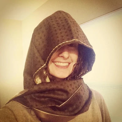 A smiling woman wearing a brown hooded scarf with a subtle pattern, partially covering her head and eyes. She is indoors, lit by warm light, and appears happy and cosy.