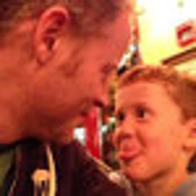 A playful moment between a man and a young boy. They are facing each other closely, smiling, with the boy sticking out his tongue. The setting appears warm and casual, likely indoors at a restaurant or café.