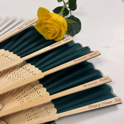 Custom engraved silk hand fans with bamboo ribs, personalised with bridal party names and wedding date, displayed with a yellow rose