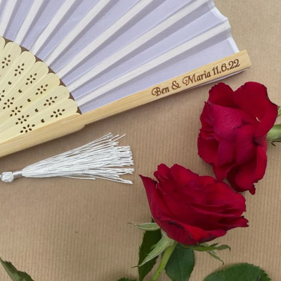 Personalised bamboo wedding hand fan with white fabric and tassel, engraved with 'Ben & Maria 11.6.22', styled with red roses.