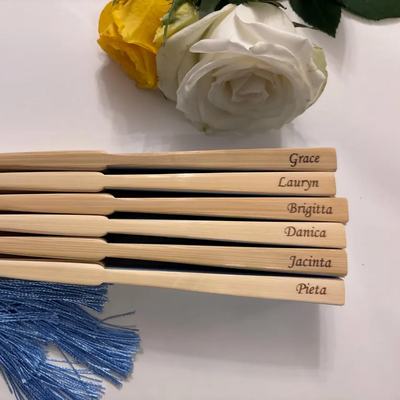 Engraved bamboo hand fans with blue tassels, personalised with names such as Grace, Lauryn, and Brigitta, styled with white and yellow roses.
