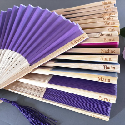 Custom engraved purple silk hand fans with bamboo ribs, featuring engraved names for wedding party gifts or keepsakes.
