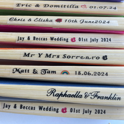 Close-up of custom printed bamboo hand fans with wedding names and dates on the main rib, including designs with hearts, roses, and rainbow icons.