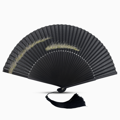 Elegant black folding hand fan with a minimalist design featuring soft, brushstroke-style grass patterns in light green and yellow tones. The fan has finely spaced black bamboo ribs, a subtle white crescent border, and a matching black tassel hanging from the base