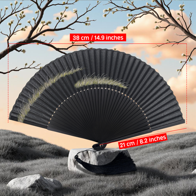 Black folding hand fan with a minimalist grass-blade design displayed outdoors on a stone. Red labels indicate dimensions: 38 cm (14.9 inches) wide when open and 21 cm (8.2 inches) tall. Background features a soft pastel sky with blooming branches and grassy terrain
