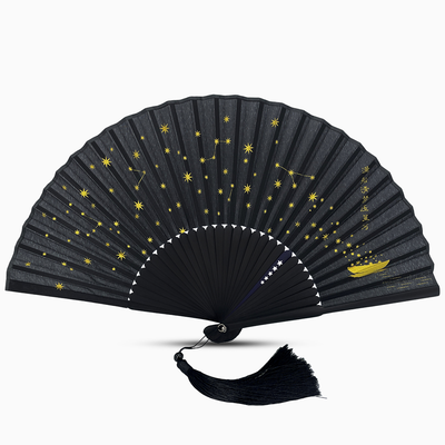 Black folding hand fan featuring a celestial design with golden stars and constellations across the fabric. A small golden boat appears on the right side with flowing star trails and vertical gold Chinese calligraphy. The fan has black bamboo ribs, a matching tassel, and a crescent white border detail along the bottom