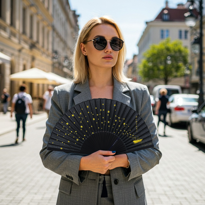 Stylish blonde woman in a grey suit and black sunglasses standing on a sunny city street, confidently holding a black folding hand fan decorated with yellow star and constellation patterns