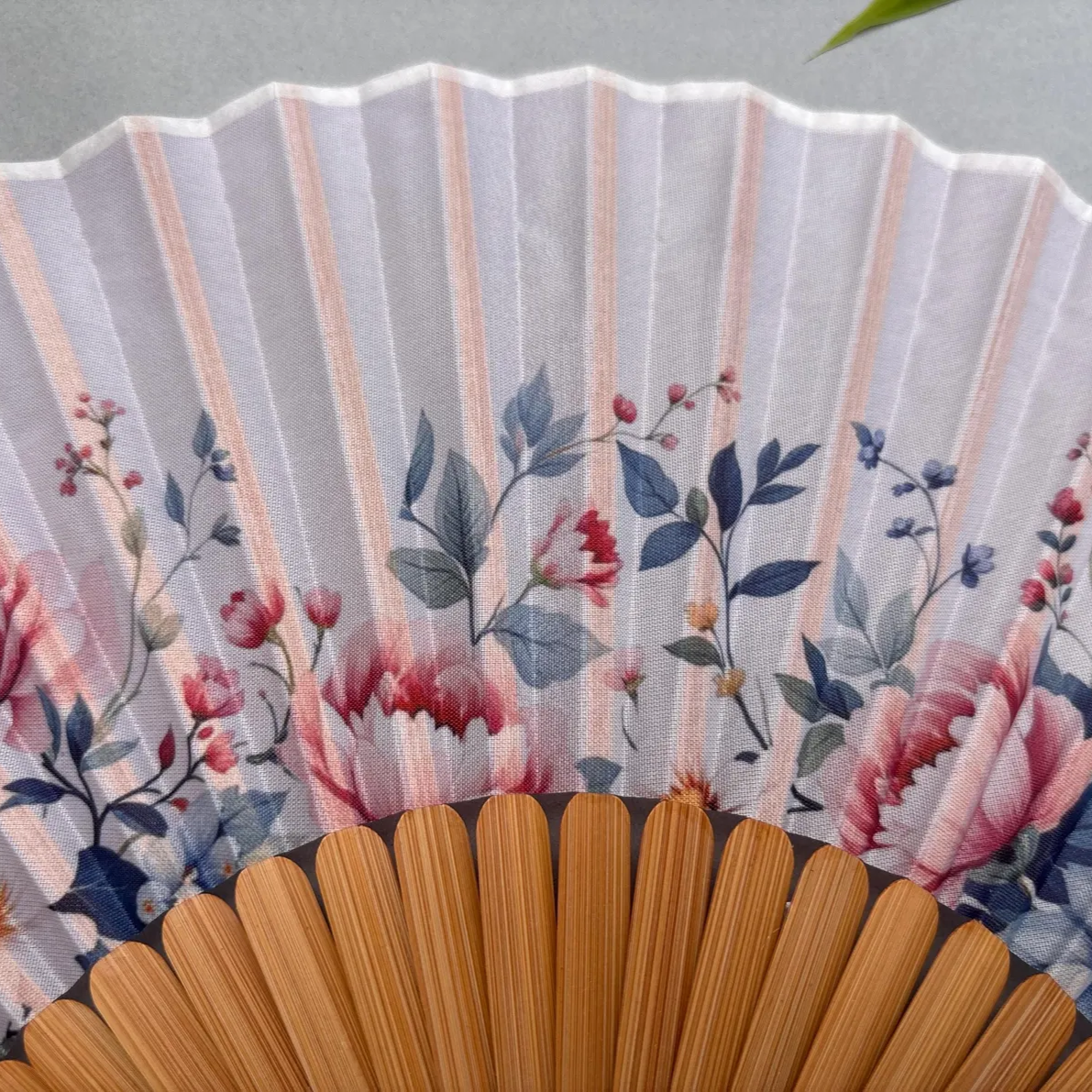 Floral-patterned hand fan with wooden frame and pink tassel on a gray background