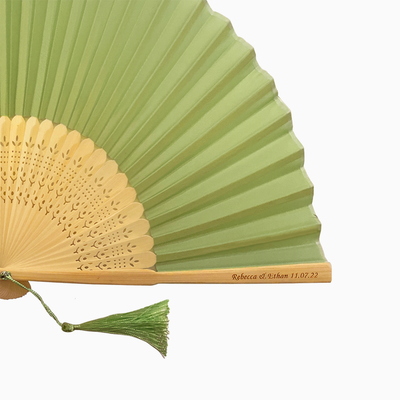 A folding fan with Sage Green and beige hues features a stylish tassel. Its engraved text on the wooden rib, radiating elegance and charm.