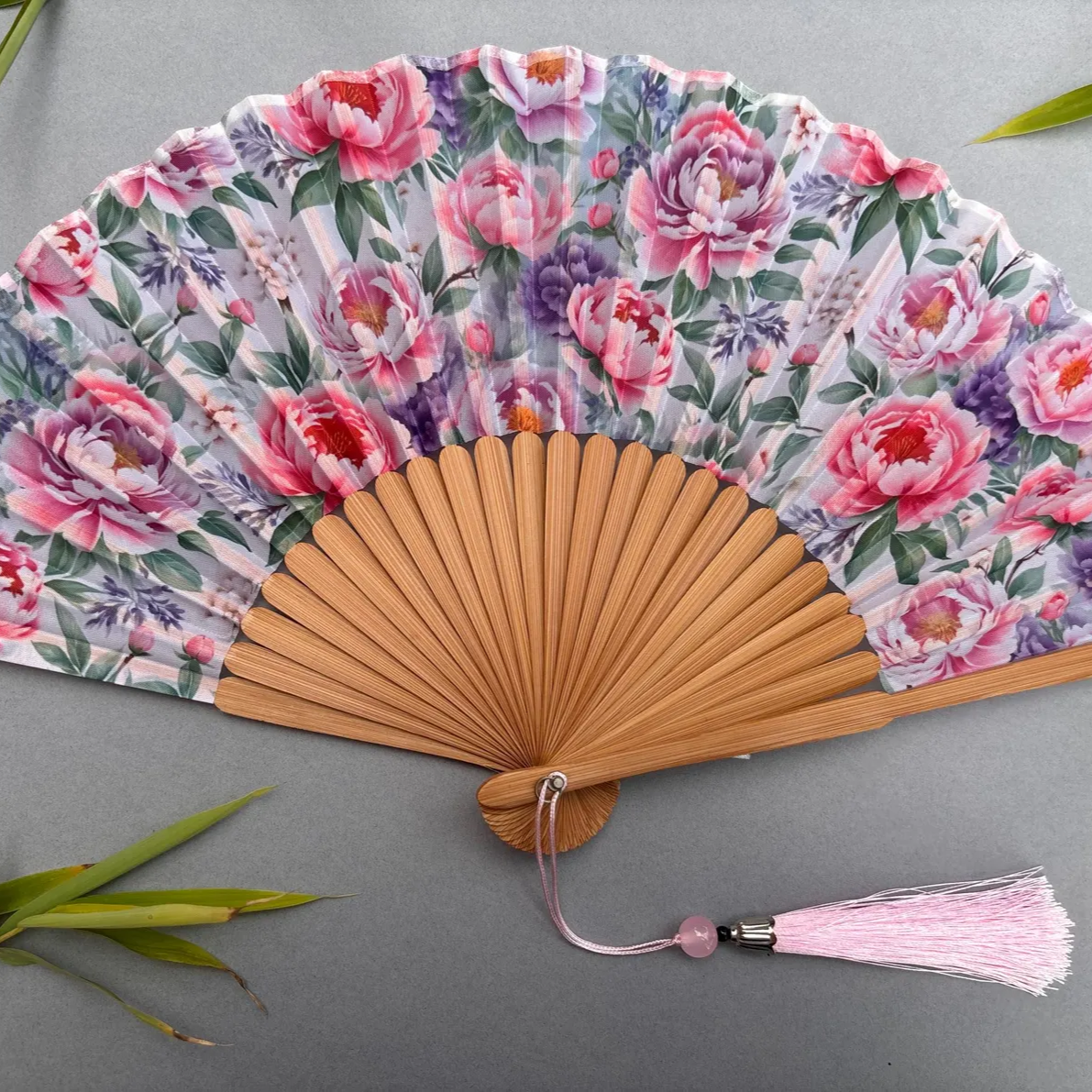 Floral-patterned hand fan with pink tassel on a gray background