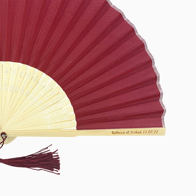 A folding fan with maroon red and beige hues features a stylish tassel. Its engraved text on the wooden rib, radiating elegance and charm.