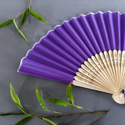 Purple folding fan with wooden handle on a gray background with green leaves.