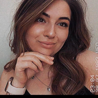 A young woman with long, wavy brown hair poses confidently with her chin resting on her hand. She is wearing a heart-shaped necklace, a smartwatch, and a sleeveless top, with a soft smile and subtle makeup. The photo has a vintage timestamp filter.